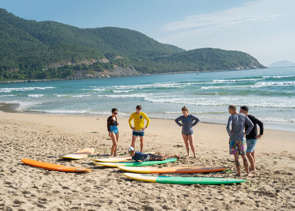 A group of five guests on Long beachat BodyVaMind Surf Yoga and Art Retreat Vietnam are gathering around a surf instructor, learning the basics of surfing before heading into the water.