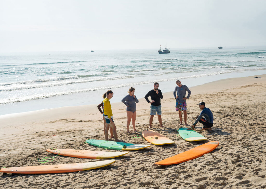 A group of people on a sandy beach in Vietnam gather around a surf instructor, learning the basics of surfing before heading into the water.