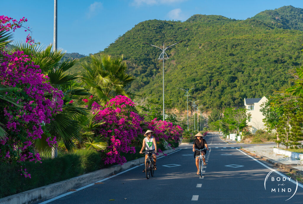 Two Spanish guests from BodyvaMind retreat riding bicycles along a scenic path in Cam Lam District, Vietnam, with the impressive Cu Hin Mountain towering in the background. Lush greenery surrounds them, capturing a moment of adventure and connection with nature