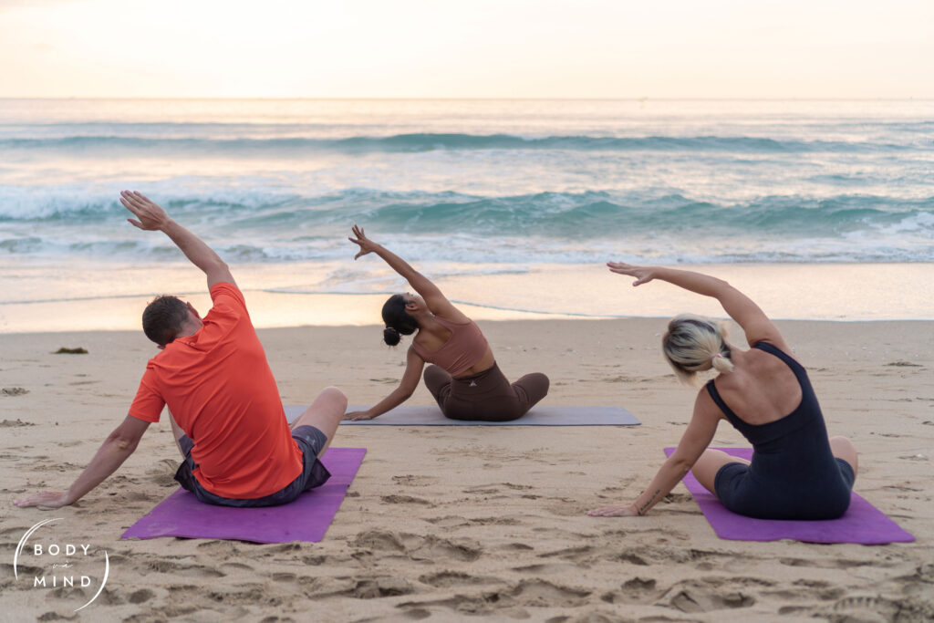 Three italian guests practicing yoga during sunrise on the beach at BodyvaMind, a surf, yoga, and art retreat in Vietnam, surrounded by golden light and ocean waves.