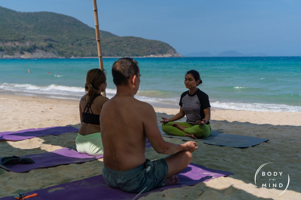 Three Vietnamese guests peacefully practice yoga at the beach at BodyvaMind, a surf, yoga, and art retreat in Vietnam, surrounded by golden light and ocean waves.