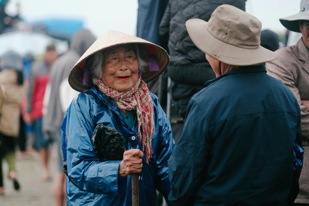 Elderly woman engaged in conversation at a bustling outdoor market, wearing a conical hat.